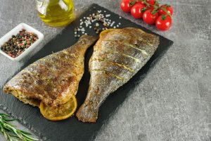 Grilled fish with roasted with lemon, rosemary, tomatoes, olive oil and spices on black slate dish, on grey background. Top view, flat lay with copy space.