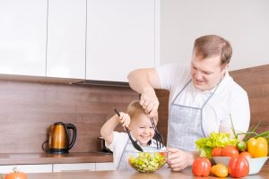 Happy family father with son preparing vegetable salad at home, vegetables and oil on the table