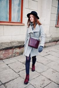 Young model girl in a gray coat and black hat with leather handbag on shoulders walking on street of city.